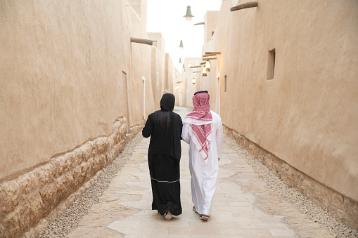 Saudi man in dish dash, kaffiyeh, and agal walking beside woman in abaya and hijab as they explore UNESCO World Heritage Site near Riyadh. Property release attached.