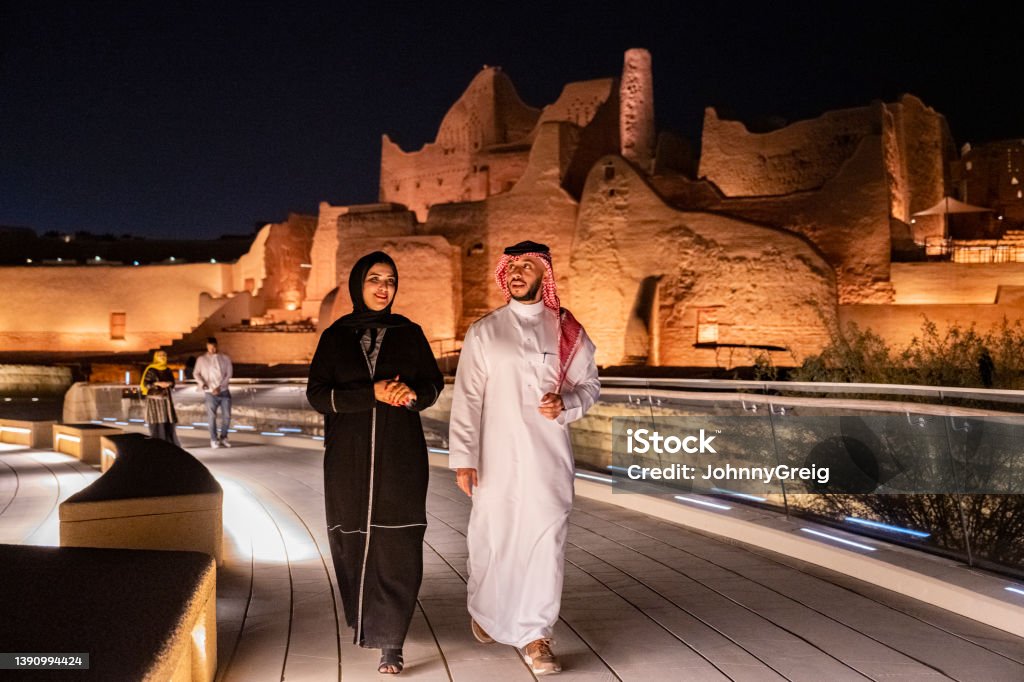 Mid adult Saudi couple exploring open air museum at night Full length view of Middle Eastern man and woman in traditional attire approaching camera as they enjoy illuminated At-Turaif ruins near Riyadh. Property release attached. Saudi Arabia Stock Photo