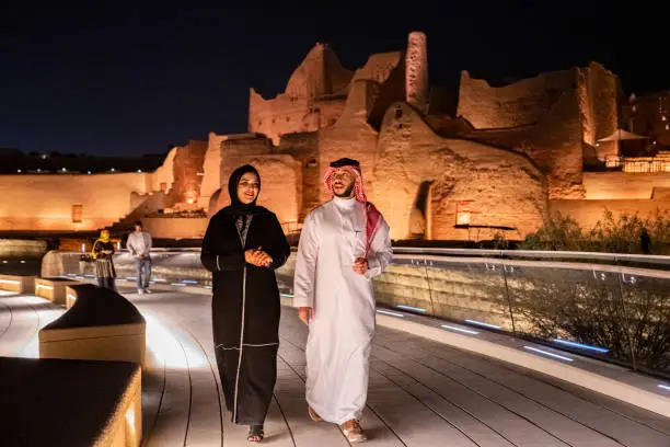 Full length view of Middle Eastern man and woman in traditional attire approaching camera as they enjoy illuminated At-Turaif ruins near Riyadh. Property release attached.