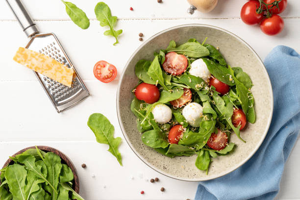 istock Fresh salad with arugula, cherry tomatoes, mozzarella cheese and hard cheese on white wooden background. Top view 1390994853