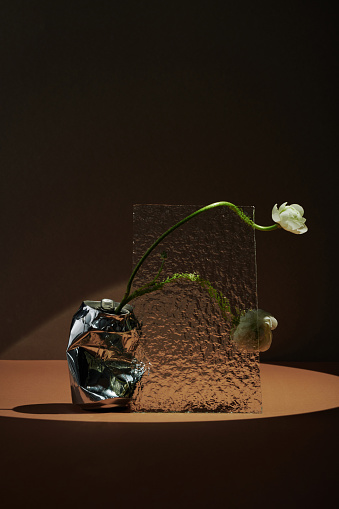 Vertical conceptual still life composition of fresh white flowers in crumpled drink can and textured glass sheet against brown background