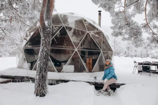 Photo of A woman sits and enjoys the view next to a dome tent in glamping in the winter forest