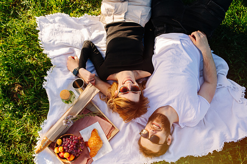 Overhead view of a young couple lying on a picnic blanket on a spring grassfield in a countryside. Wearing sunglasses, smiling.