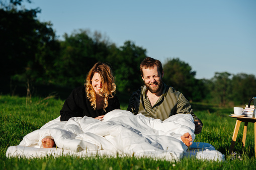 Couple waking up from a sweet long nap in bed made outside on a grass in a countryside. Both groggy and happy. Bare feet sticking from under a blanket.