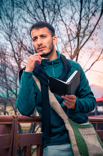 In this outdoor dusk image, an Asian/Indian young adult male writer/poet contemplates deeply by holding a diary and pen while engrossed in writing. He carries a shoulder bag and wears a warm sweater and neck scarf. He writes in the hills of Himachal Pradesh.