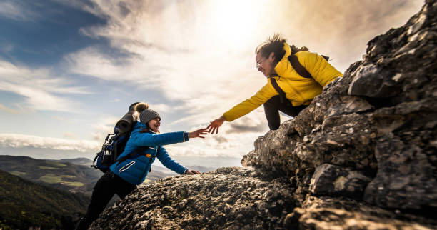 People helping each other hike up a mountain at sunrise - Giving helping hand and teamwork concept stock photo