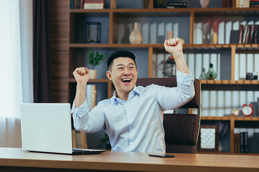 Happy businessman dancing and celebrating victory sitting at a table in a classic office, Asian working at computer