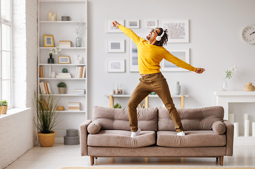 Full body black man in casual clothes and wireless headphones raising arms, jumping and dancing on sofa while having fun in living room in weekend at home