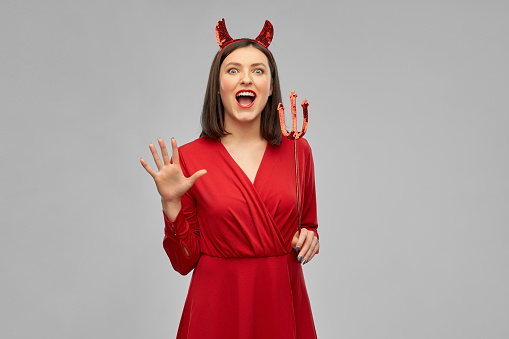 holiday, theme party and people concept - scary woman in red halloween costume of devil with horns and trident over grey background
