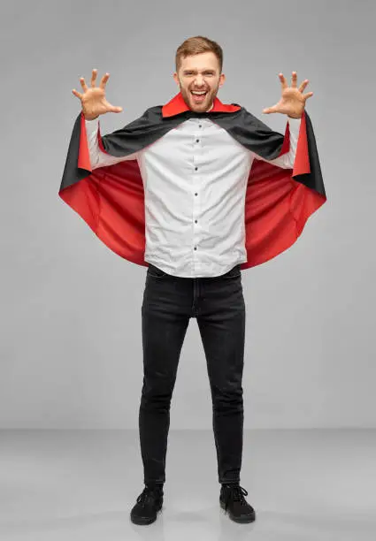 holiday, theme party and people concept - man in halloween costume of vampire and dracula cape scaring over grey background