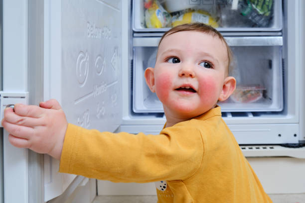 el bebé pequeño con alergias en las mejillas rojas se subió a un refrigerador abierto. problemas de seguridad infantil en la habitación del hogar, niño pequeño - mejillas enrojecidas fotografías e imágenes de stock