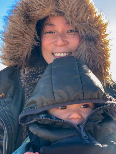 A selfie shot of a woman with her baby boy on her lap outdoors in Northumberland, England. They are wearing winter coats during a cold season. She is smiling while looking at the camera.
