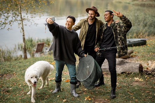 Joyful multiracial male friends taking selfie on smartphone during resting in nature. Men fishing and spending time together on river or lake coast at autumn. Leisure, weekend and vacation. Friendship