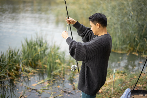 Asian man fishing on river or lake coast alone. Concept of leisure, hobby and weekend in nature. Autumn day