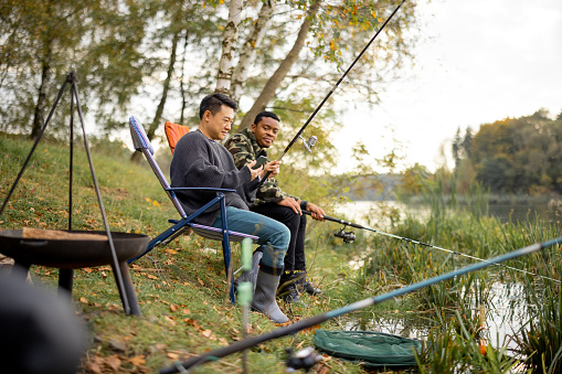 Two multiracial male friends fishing in nature. Men resting and spending time together on river or lake shore. Concept of leisure, hobby and weekend in nature. Friendship