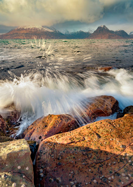 Crashing Waves At Elgol, Isle of Skye, UK. Crashing waves on a cold Winter afternoon at Elgol on the Isle of Skye, Scotland. Sunlight can be seen in the foreground rocks with a dark and moody sky and snowcapped mountains. elgol beach stock pictures, royalty-free photos & images