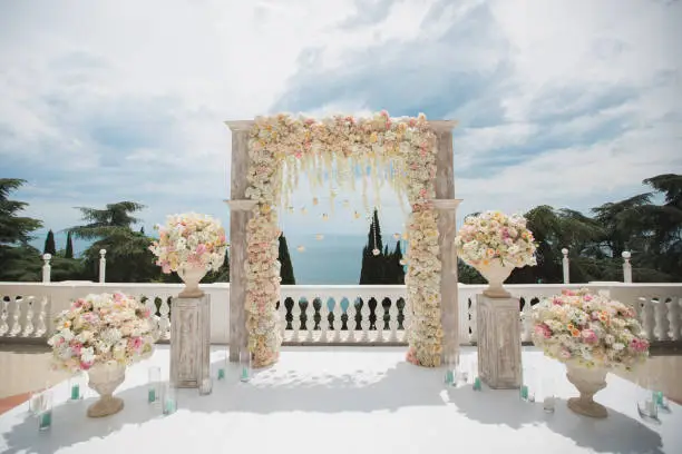 Photo of Elegant wedding arch with fresh flowers, vases on background of ocean and blue sky