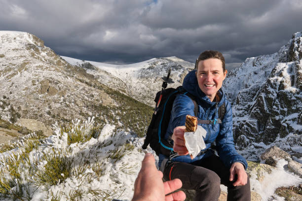 Content female hiker with backpack sitting in snowy mountains and looking at camera while suggesting chocolate bar in overcast weather