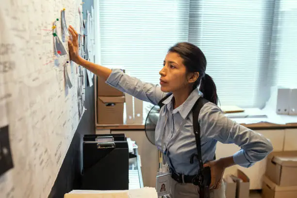 Young serious FBI agent in uniform looking at map with pinned photos of criminals in office while thinking of ideas of their location