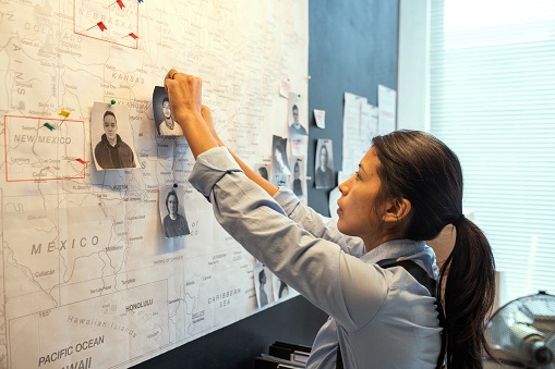 Young Hispanic female agent of FBI in uniform pinning photos of several male suspects on map while suggesting location of their gang