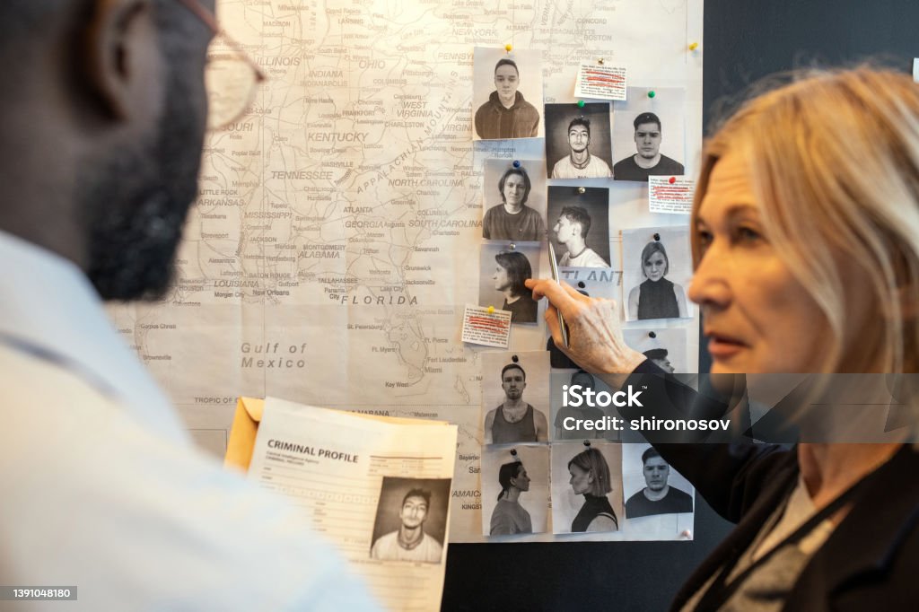 Blond confident female detective pointing at photo of suspect on board Blond confident female detective pointing at photo of suspect on board over map while matching facts of serial crimes with colleague Criminal Investigation Stock Photo