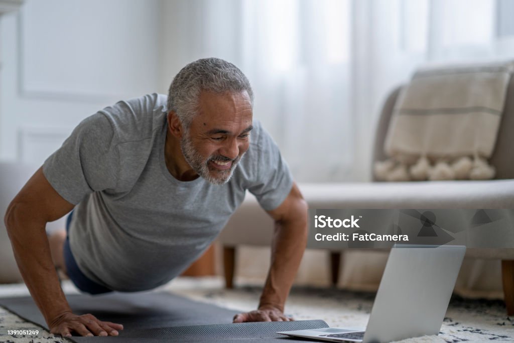 Senior Gentleman Exercising Virtually at Home A senior gentleman of African decent, lays out on a yoga mat in his living room as he exercises along with a virtual class.  He is dressed comfortably in athletic wear and has his laptop out in front of him as he follows along with the instructor and does push-ups. Exercising Stock Photo