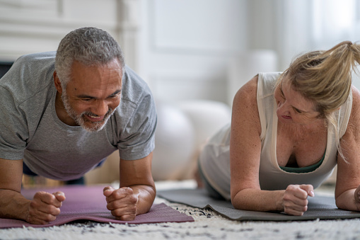 A senior couple lay out on yoga mats in their living room as they practice some strength conditioning at home together.  They are both dressed comfortably in  athletic wear and are holding a planking position.