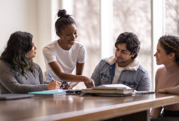 University Students Helping Each Other A young female University student of African decent, stands behind a peer and leans in as she tries to help her classmate with his studies. They are both dressed casually and are among a group sitting at the table all studying together for class. group of students stock pictures, royalty-free photos & images