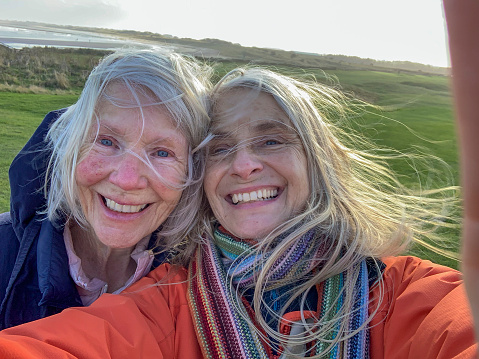 A selfie shot of a woman and her daughter outdoors in Northumberland, England with the coastline behind them. They are looking at the camera and smiling while the wind is blowing their hair around.