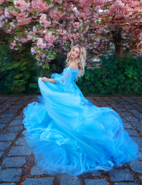 princesse femme heureuse fantastique dans une longue robe bleue moelleuse, comme cendrillon, tourne en rond dans la danse, le tissu vole des vagues en mouvement. fée fille déesse cheveux blonds. nature à floraison printanière, fleurs roses de l’arbre - women nymph hairstyle mythology photos et images de collection