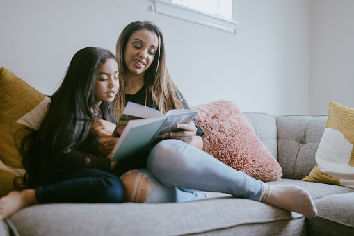 A multiracial mom and her girl snuggle together on the living room sofa, reading a picture books together.  Reading to children increases intelligence and connection.