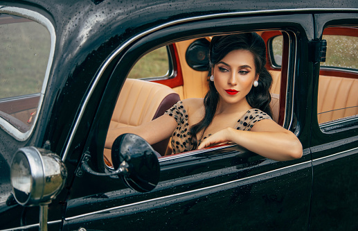 Elegant young retro woman sits behind wheel and looks out of window. Portrait elegance Beautiful girl driver in vintage dress, pin-up hairstyle, sexy eyes, face with red lips makeup. Bad weather rain.