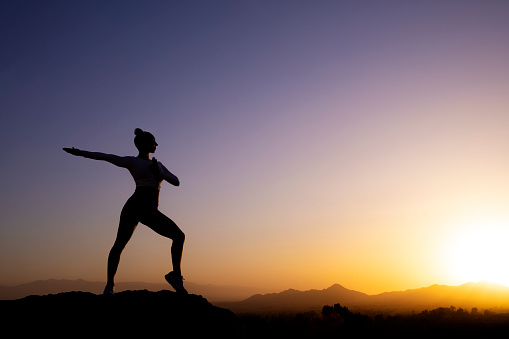 This is a conceptual photo showing a silhouette woman doing yoga and exercise outdoors on the rocks at sunset