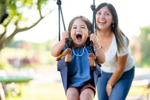A single Mom pushes her daughter on a swing while spending some quality time together outside. The little girl is dressed casually and wearing a hat to keep the warm summer sun off her face. She is giggling with each push her mother gives her.