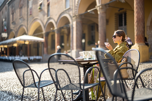 Stylish woman sitting at outdoor cafe in the old town of Bologna city. Italian measured lifestyle and street fashion concept. Idea of traveling Italy