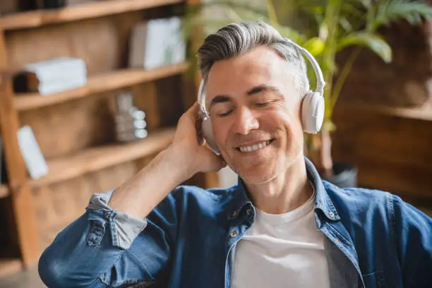 Relaxed caucasian mature middle-aged man freelancer listening to the music song playlist radio podcast in headphones earphones in mobile application online at home office