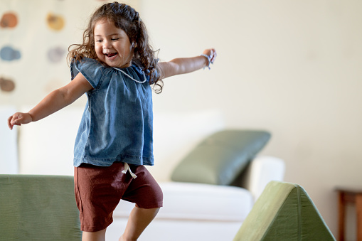 A sweet little girl of Hispanic decent, dances joyfully around her living room all by herself.  She is dressed casually in a blue top and burgundy shorts as she stretches her arms out and twirls around the room with glee.