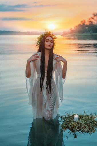 Photo with noise. Pagan girl Slavic goddess emerges from water lake, ritual flower wreaths float, candles burning. fashion model. Fantasy woman enjoy summer sunset. Wet white vintage medieval dress