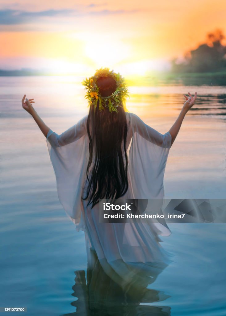 Photo with noise. Fantasy woman standing in water hands raised to sky, praying to sun. Slavic girl in herbal wreath, long hair white dress, pagan divination ritual. Nature blue river sunset. Back view Women Stock Photo
