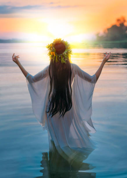 photo with noise. fantasy woman standing in water hands raised to sky, praying to sun. slavic girl in herbal wreath, long hair white dress, pagan divination ritual. nature blue river sunset. back view - witch beauty beautiful women imagens e fotografias de stock