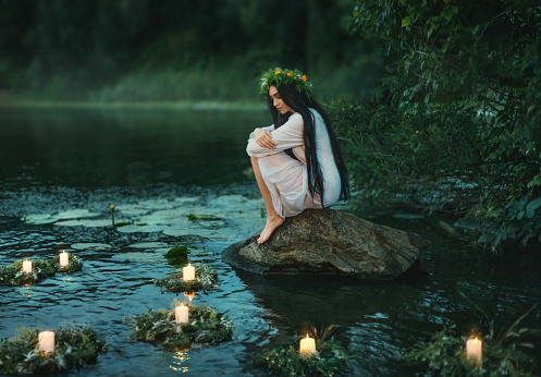 Slavic girl sits on stone on shore lake. Nymph fantasy woman hugs knees. Long black hair. Wreaths of grass, flowers float on water. Candles burning. River dusk forest green tree. Holiday Ivan Kupala.