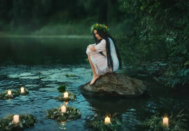 Slavic girl sits on stone on shore lake. Nymph fantasy woman hugs knees. Long black hair. Wreaths of grass, flowers float on water. Candles burning. River dusk forest green tree. Riutal of Divination