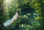 photo with noise. mythical natural landscape. Forest fantasy woman sits on shore lake, nymph throws wreath in water. Candles burning. Old Slavic cult ceremony Ivan Kupala. Long white dress. Black hair