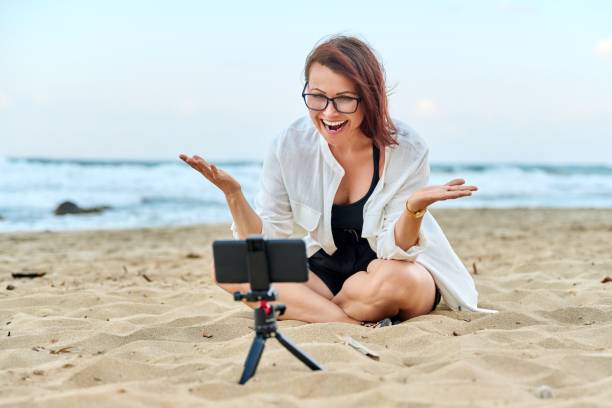 istock Middle aged woman sitting on beach with smartphone using video call 1391075339