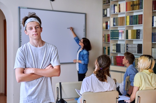 Teenage guy in classroom looking at camera, group of high school students with teacher study in library. Education, learning, knowledge, adolescence concept