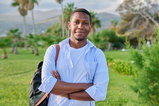Outdoor portrait of African American handsome man with crossed arms looking at camera. Positive confident 30s male with backpack, tropical park nature background.