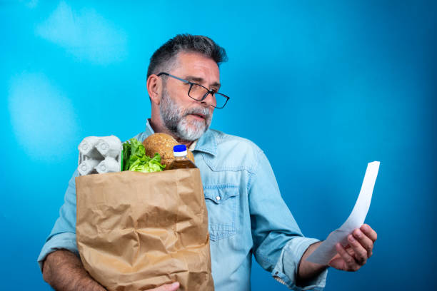 Shocked mature man looking at store receipt after shopping, holding a paper bag with healthy food. Real people expression. Inflation concept. man with a paper bag of groceries looks surprised. hombre maduro conmocionado mirando el recibo de la tienda después de comprar, sosteniendo una bolsa de papel con alimentos saludables. expresión de personas reales. concepto de inflación. hombre con una bolsa de papel de comestibles parece sorprendido.