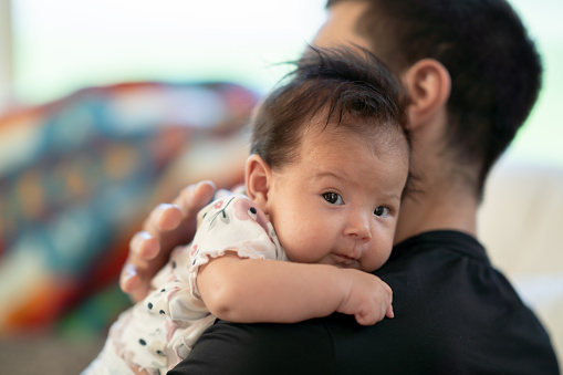 A sweet little newborn looks at the camera as she is propped up on her fathers shoulder. The sweet little indigenous baby has dark fluffy hair and is nestling into her fathers neck and shoulder.