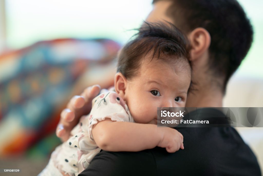 Bonding Time with Daddy A sweet little newborn looks at the camera as she is propped up on her fathers shoulder. The sweet little indigenous baby has dark fluffy hair and is nestling into her fathers neck and shoulder. Baby - Human Age Stock Photo