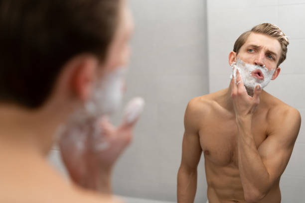 serious man with shaving foam on face in front of mirror, morning of young caucasian male in bathroom, hygiene and skin care - shaving men shaving cream mirror imagens e fotografias de stock